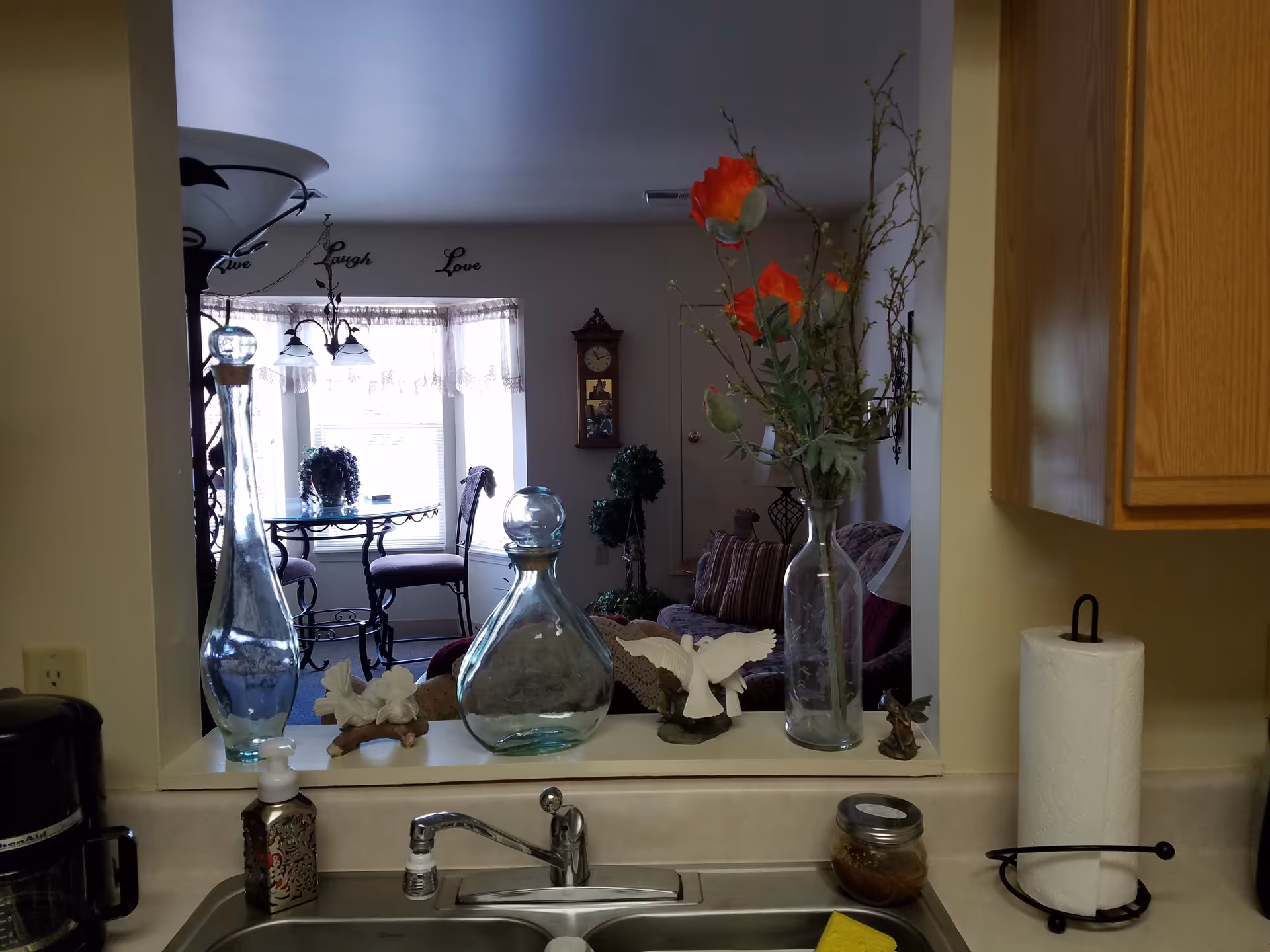 View from a kitchen sink through a pass-through into a living/dining area with decorative glass vases, flowers, and a table by a bay window.