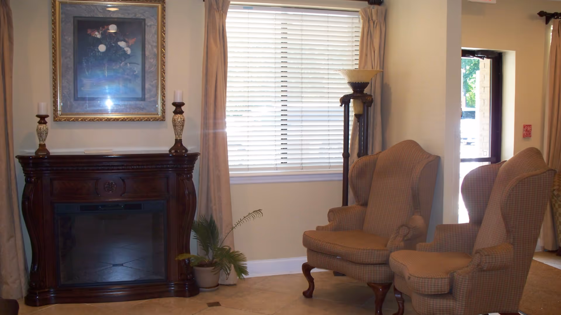 A cozy interior room featuring two upholstered wingback chairs, a wooden electric fireplace with decorative candles on top, a framed painting above the fireplace, a tall floor lamp, a potted plant, and a window with closed blinds and beige curtains. There is also a door leading outside visible on the right side.
