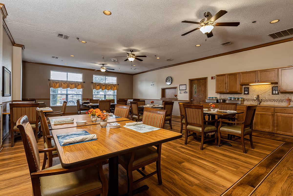 A spacious dining room with wooden tables and chairs arranged neatly. Each table has placemats, napkins, and small flower vases. The room features large windows with valance curtains, ceiling fans with lights, and wooden cabinetry along one wall with a countertop and sink area.