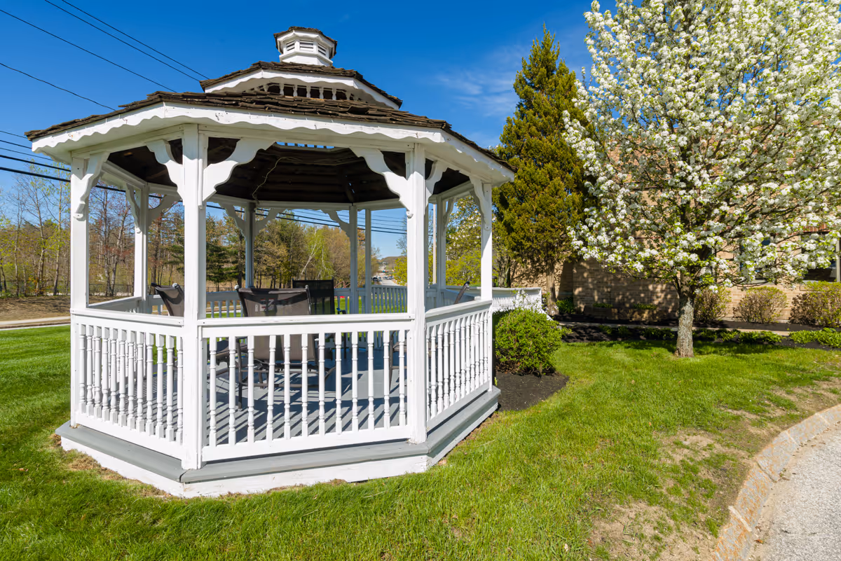 A white wooden gazebo with a shingled roof situated on a green lawn. There are chairs inside the gazebo. To the right, a tree with white blossoms and some bushes are visible. The sky is clear and blue.
