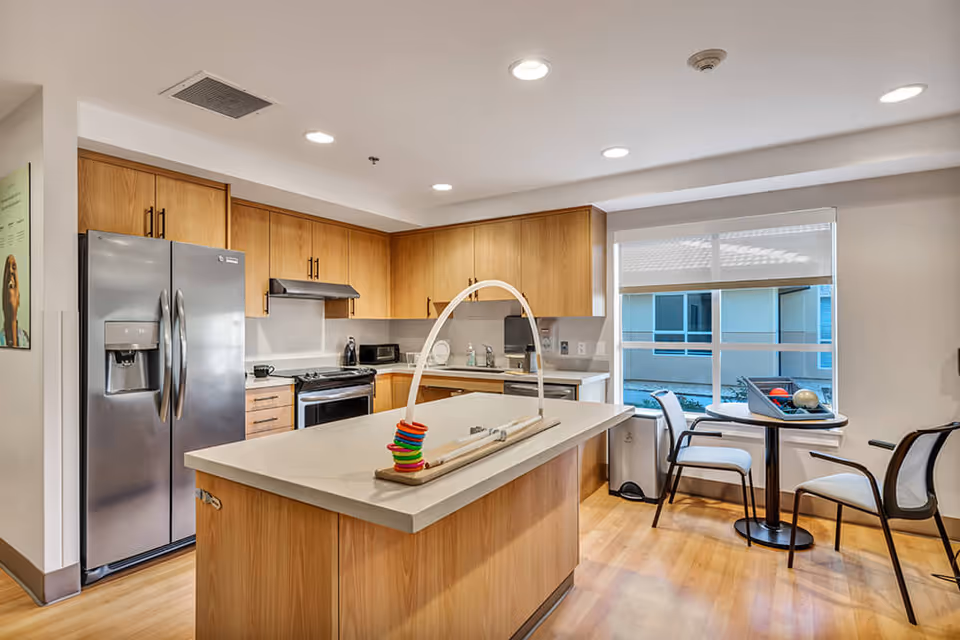 A modern kitchen area with wooden cabinets, a stainless steel refrigerator, stove, and dishwasher. There is a kitchen island in the center with a colorful ring stacking toy on top. To the right, there is a small round table with three chairs near a window that looks out onto an outdoor courtyard.