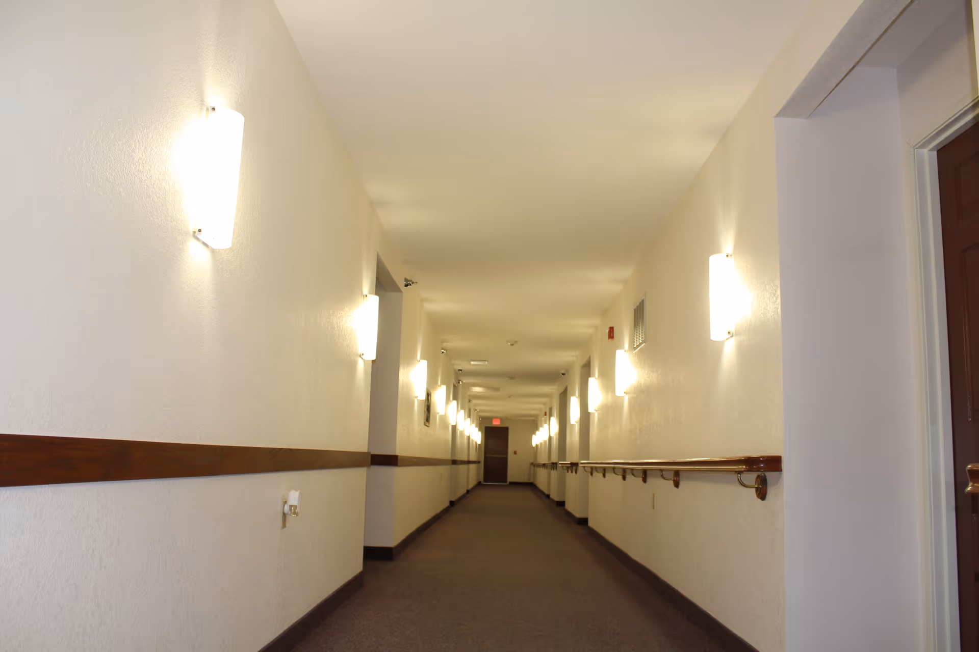 A long, well-lit interior hallway with wall-mounted lights and wooden handrails leading to a door at the far end.
