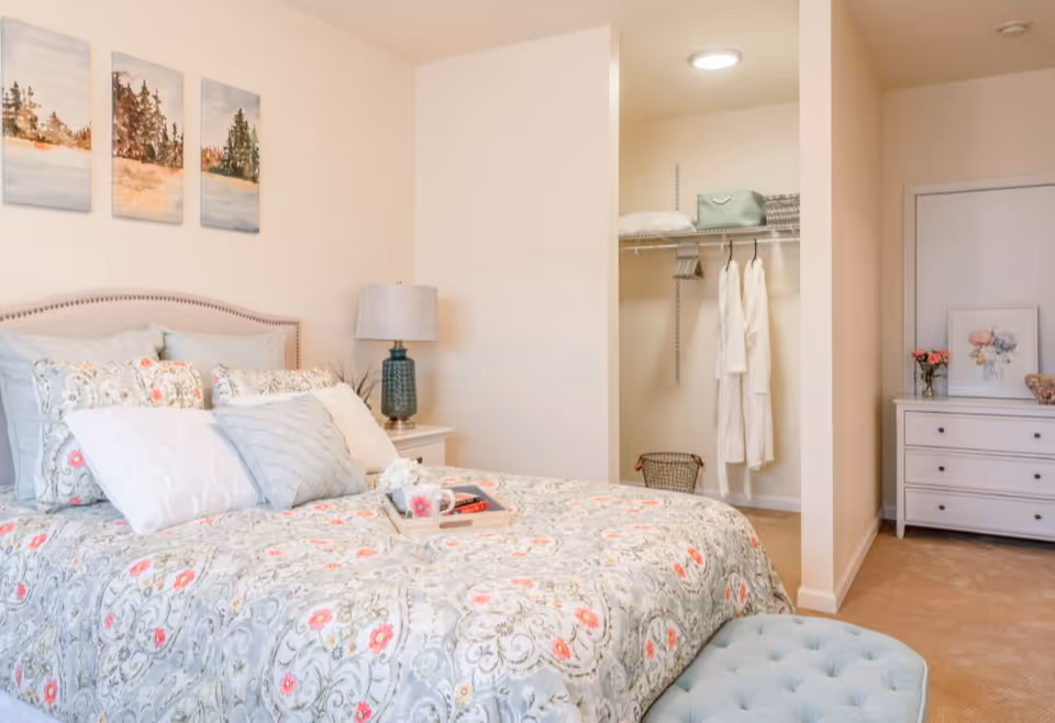 A cozy bedroom with a neatly made bed featuring floral patterned bedding and multiple pillows. A tray with a cup and a book rests on the bed. To the left of the bed is a nightstand with a green lamp. On the wall above the bed are three landscape paintings. To the right, there is an open closet with hanging robes and shelves holding pillows and storage baskets. A white dresser with flowers and framed artwork sits against the far wall.