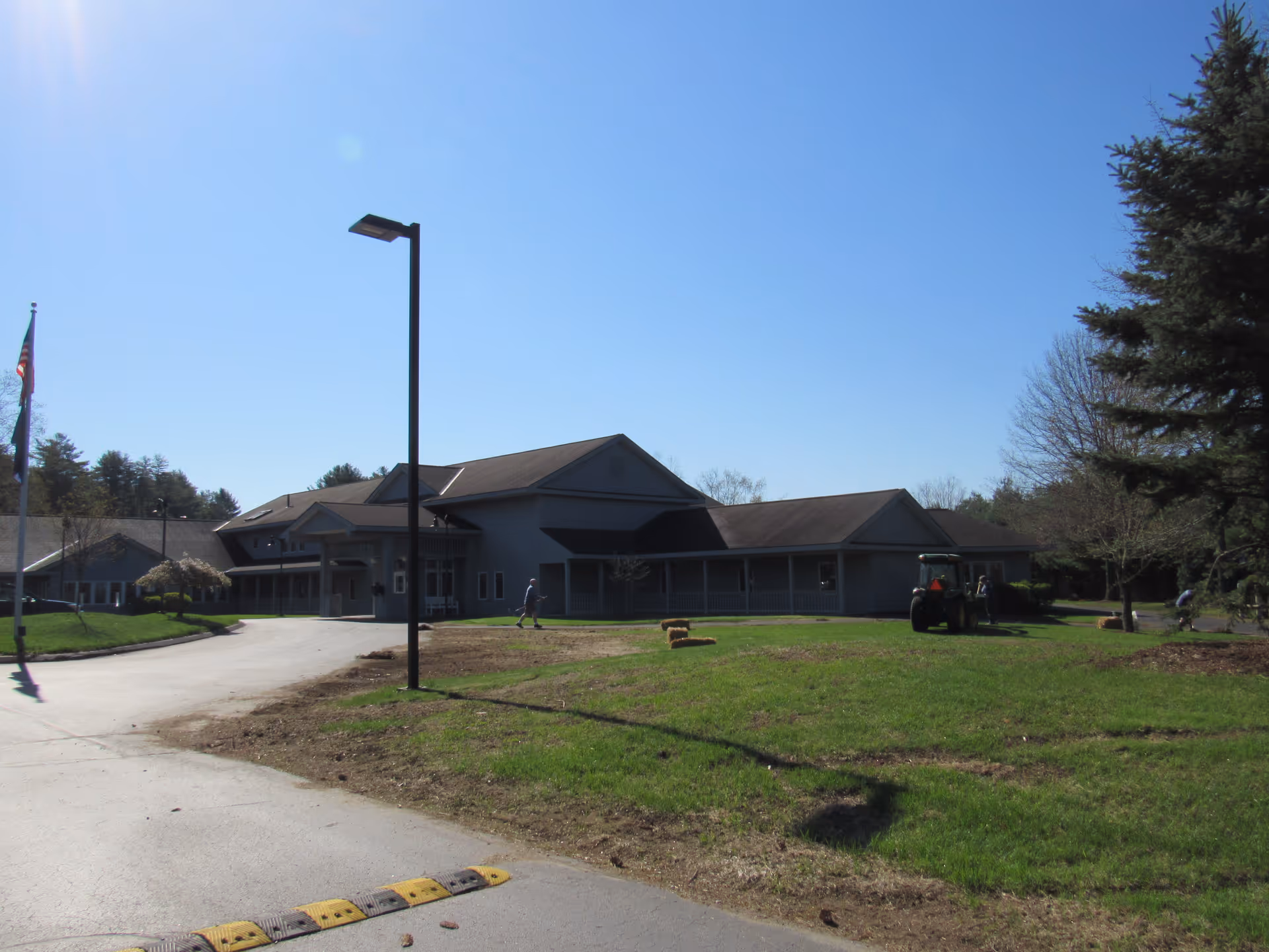 Single-story institutional building with a driveway, lamp post, grassy lawn and a tractor under a clear blue sky.