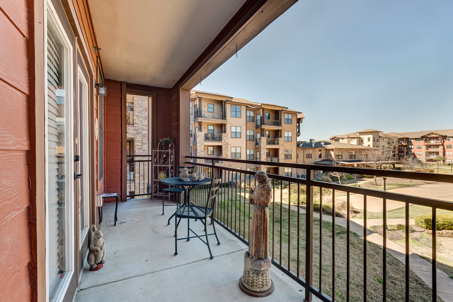 A covered balcony with a small round table and two chairs, decorative statues including a rabbit and a figure, overlooking a courtyard with grass and pathways, surrounded by multi-story residential buildings under a clear blue sky.