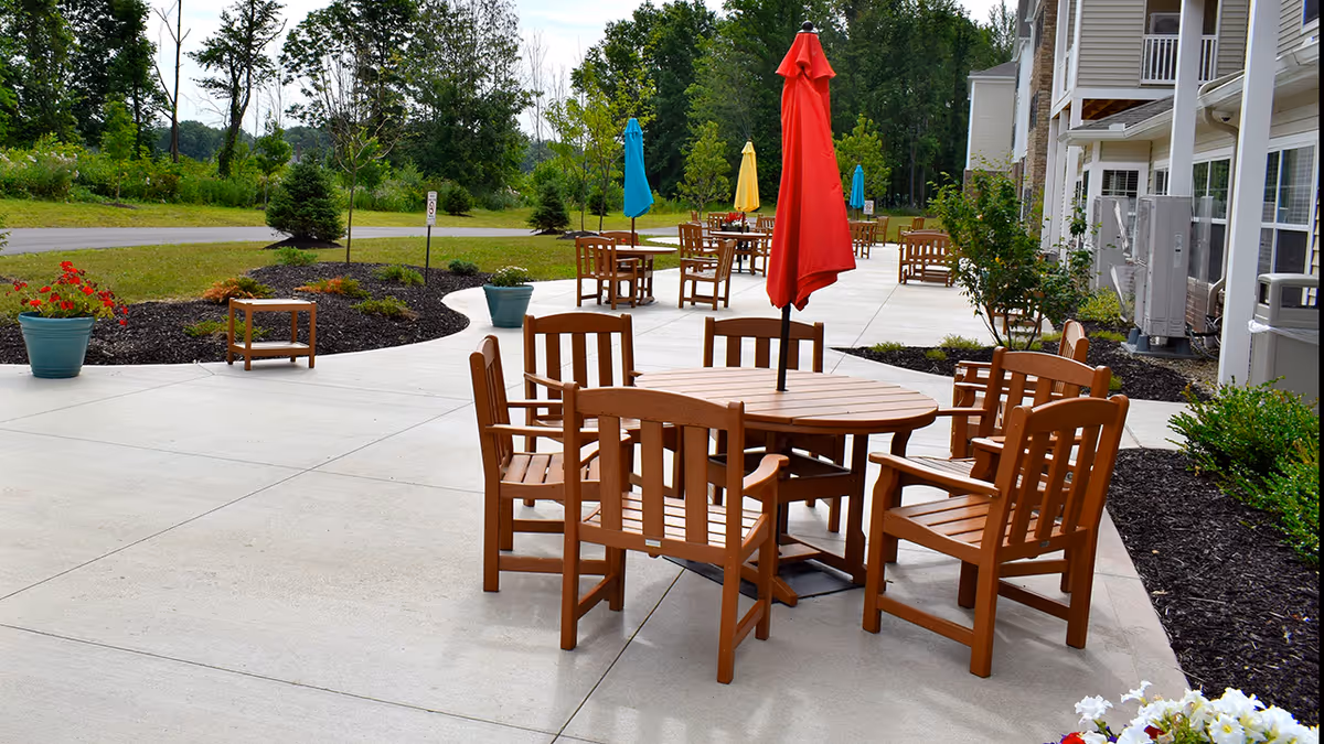 Outdoor patio area with multiple round wooden tables and matching chairs. Each table has a colorful closed umbrella in red, blue, or yellow. The patio is surrounded by landscaped greenery and trees, with a building visible on the right side.
