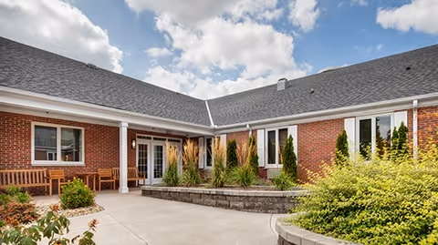 Outdoor courtyard area of a senior living facility with brick walls, multiple windows, a concrete walkway, wooden benches, and landscaped plants including tall grasses and shrubs under a partly cloudy sky.