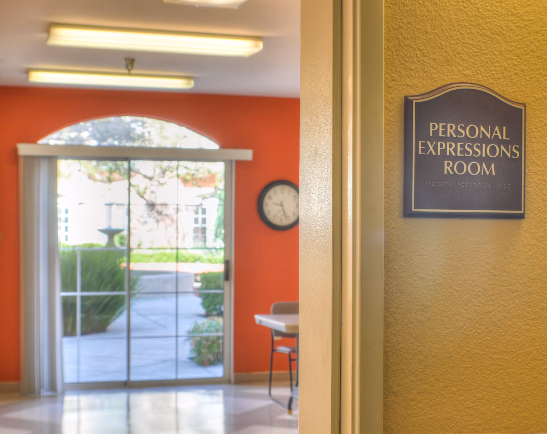 Interior view of a room entrance with a sign that reads 'Personal Expressions Room' on a textured yellow wall. In the background, there is a sliding glass door with vertical blinds, an orange wall, a clock, and a table with chairs. Outside the glass door, greenery and a fountain are visible.