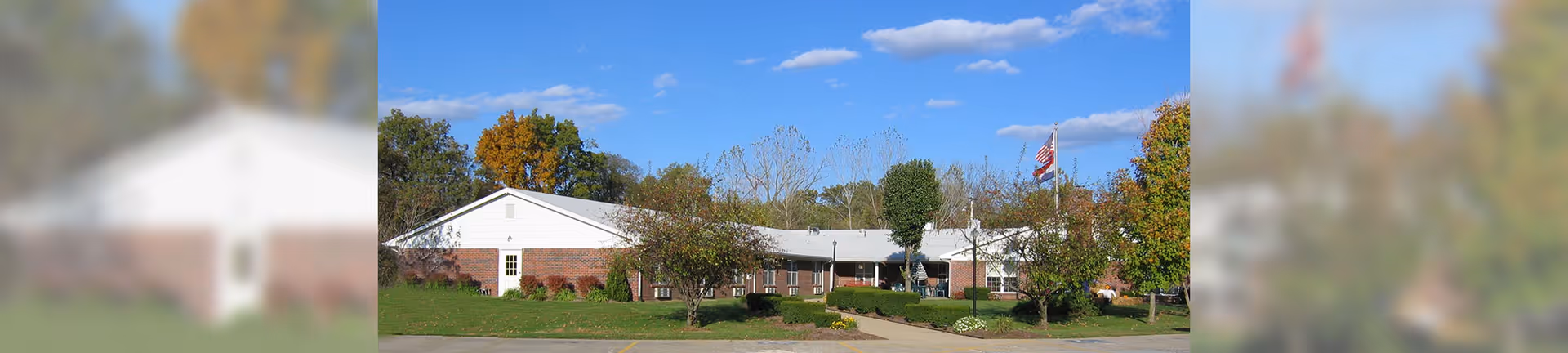 Exterior view of St Genevieve Care Center, a single-story brick building with a white roof, surrounded by trees and greenery under a blue sky with scattered clouds. There is a pathway leading to the entrance and two flagpoles with flags flying.