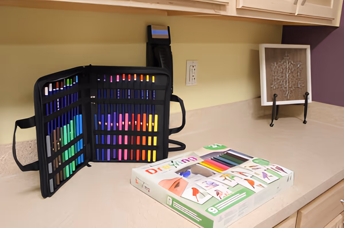 A countertop with an open case of colorful markers or pens arranged neatly inside, next to a box labeled 'Drawing' containing colored pencils or markers. In the background, there is a framed piece of artwork on a stand against a wall with light yellow and purple paint.