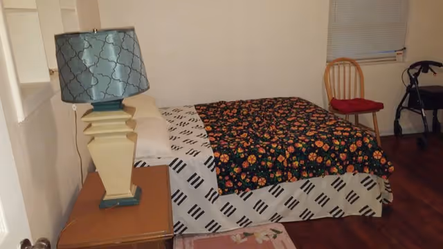 A furnished bedroom with a bed, lamp, nightstand, and a walker by the window.