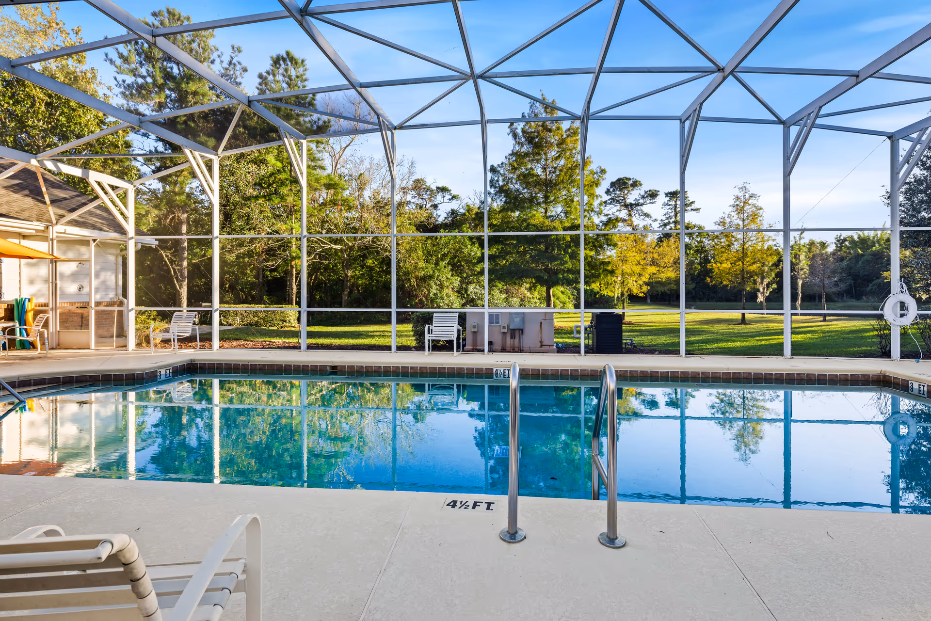 An outdoor swimming pool enclosed by a white metal and screen structure, surrounded by green trees and grass under a clear blue sky. There are white poolside chairs and a life preserver attached to the enclosure.