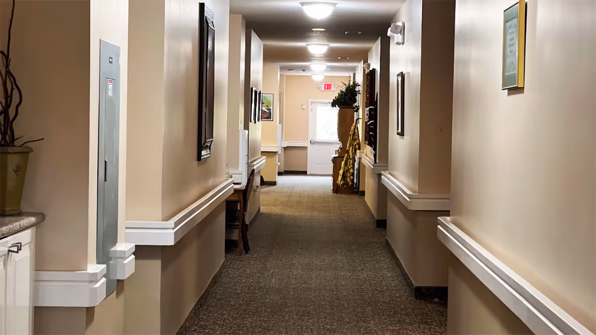A well-lit indoor hallway with beige walls and carpeted floor, featuring handrails on both sides, framed pictures on the walls, a small table with chairs on the left side, and a door with a window at the far end.