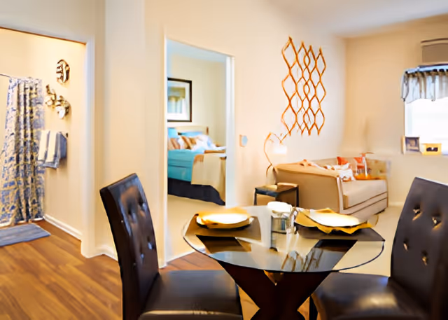Interior view of an assisted living facility showing a small dining area with a round glass table set for two with yellow plates and dark cushioned chairs. In the background, there is a living room area with a beige sofa, a decorative wall piece, and a window with a valance. To the left, a doorway leads to a bedroom with a bed and framed artwork, and another doorway leads to a bathroom with a blue patterned shower curtain and towels.