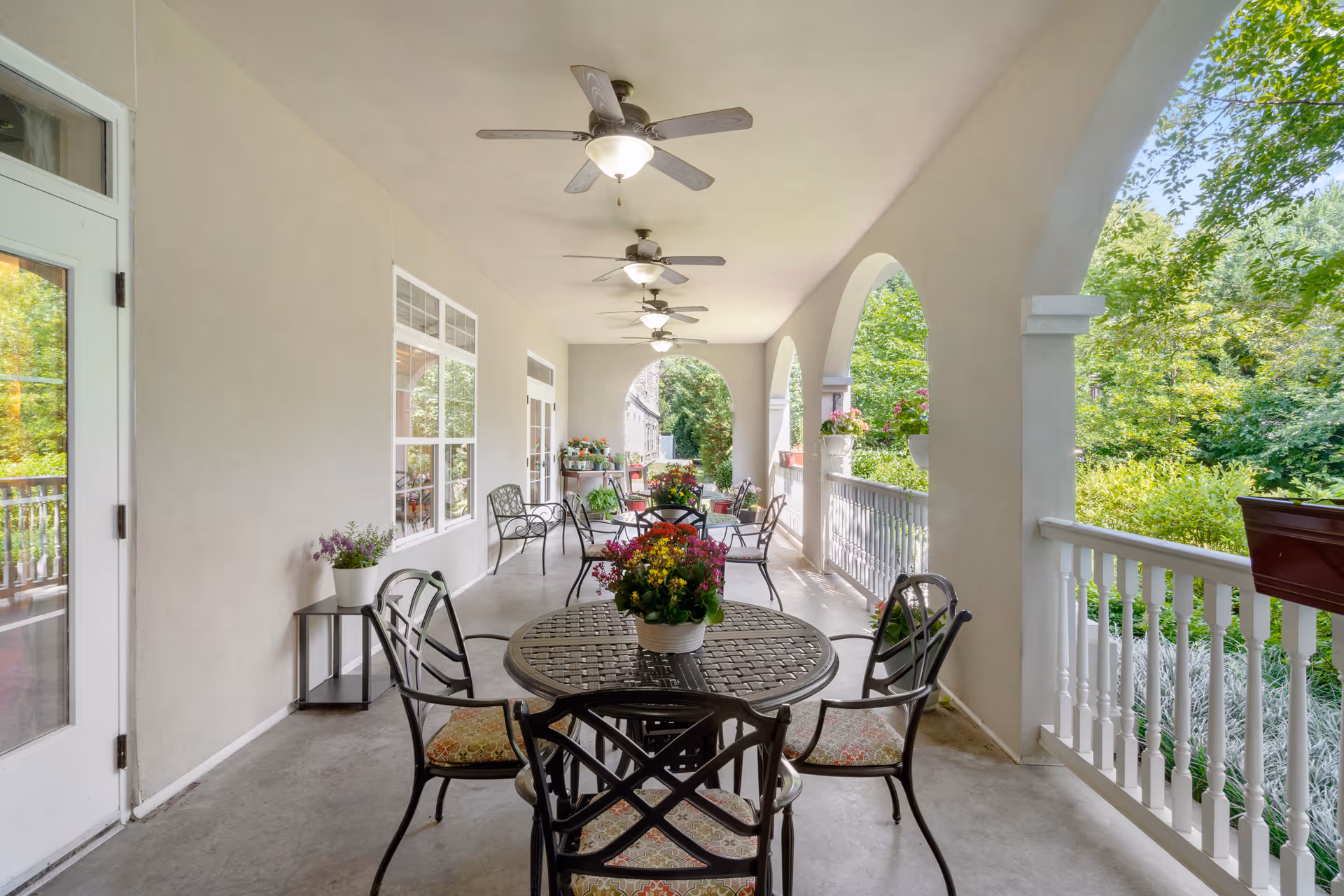 Covered outdoor porch with round tables and metal chairs, ceiling fans, and potted flowers overlooking green landscaping.