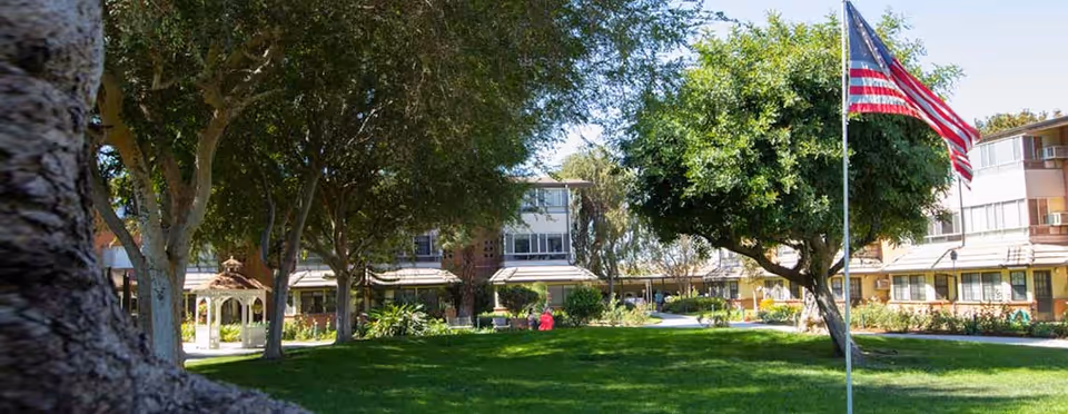 A well-maintained outdoor garden area at Rowntree Gardens with lush green grass, several large trees providing shade, a gazebo, and an American flag on a flagpole. The background shows a multi-story residential building with windows and balconies.