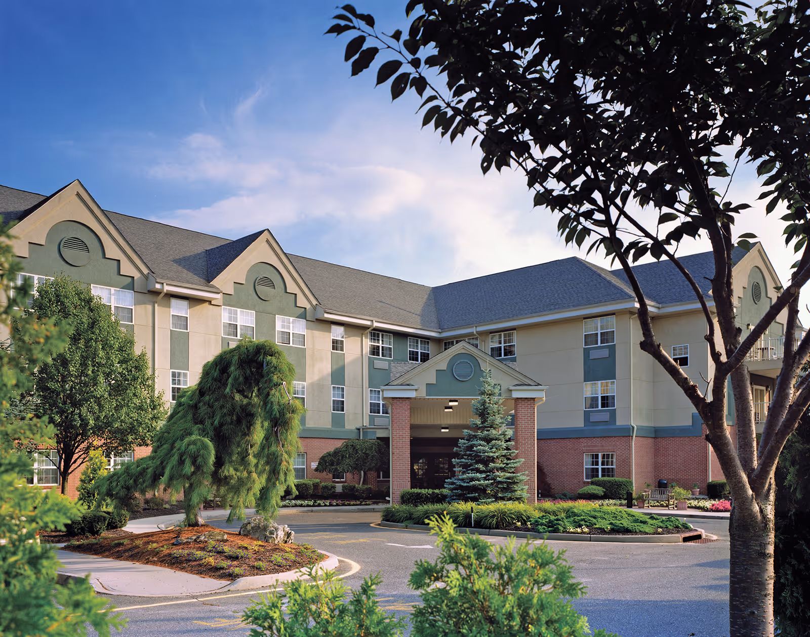 Front exterior of a multi-story senior living facility with a covered entrance, landscaped driveway, and trees.