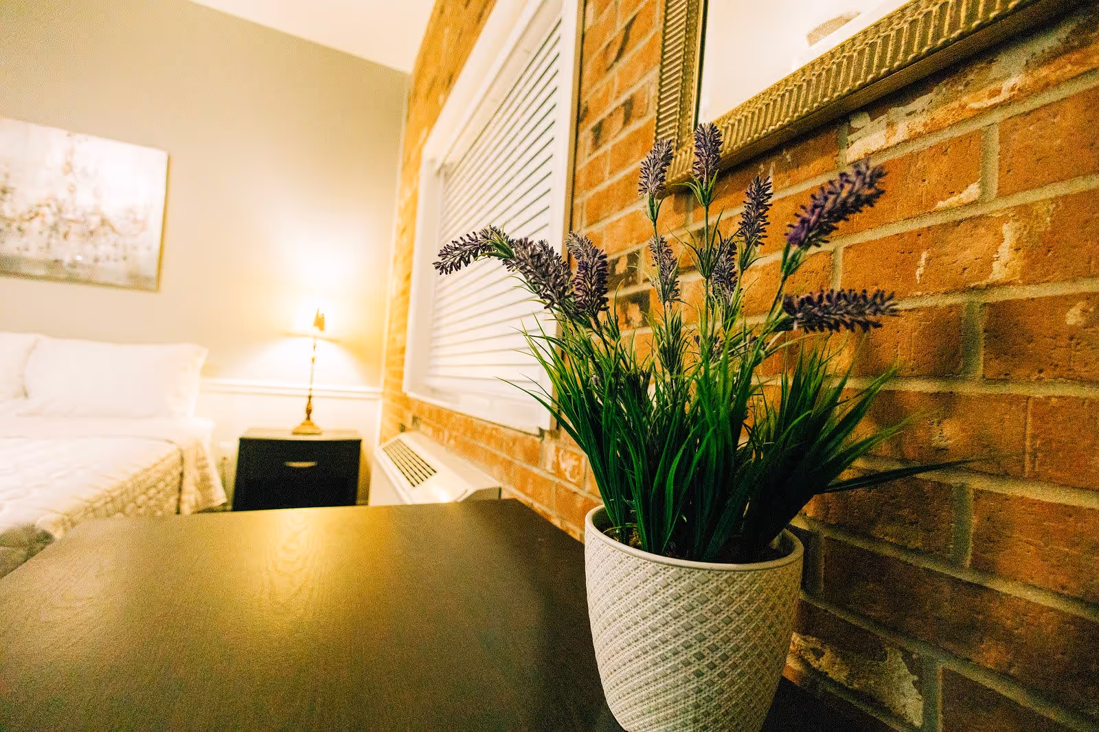 A cozy bedroom corner featuring a potted plant with purple flowers on a dark wooden surface, a brick wall with a framed mirror, a window with closed blinds, and a bedside table with a lamp and a bed with white bedding in the background.