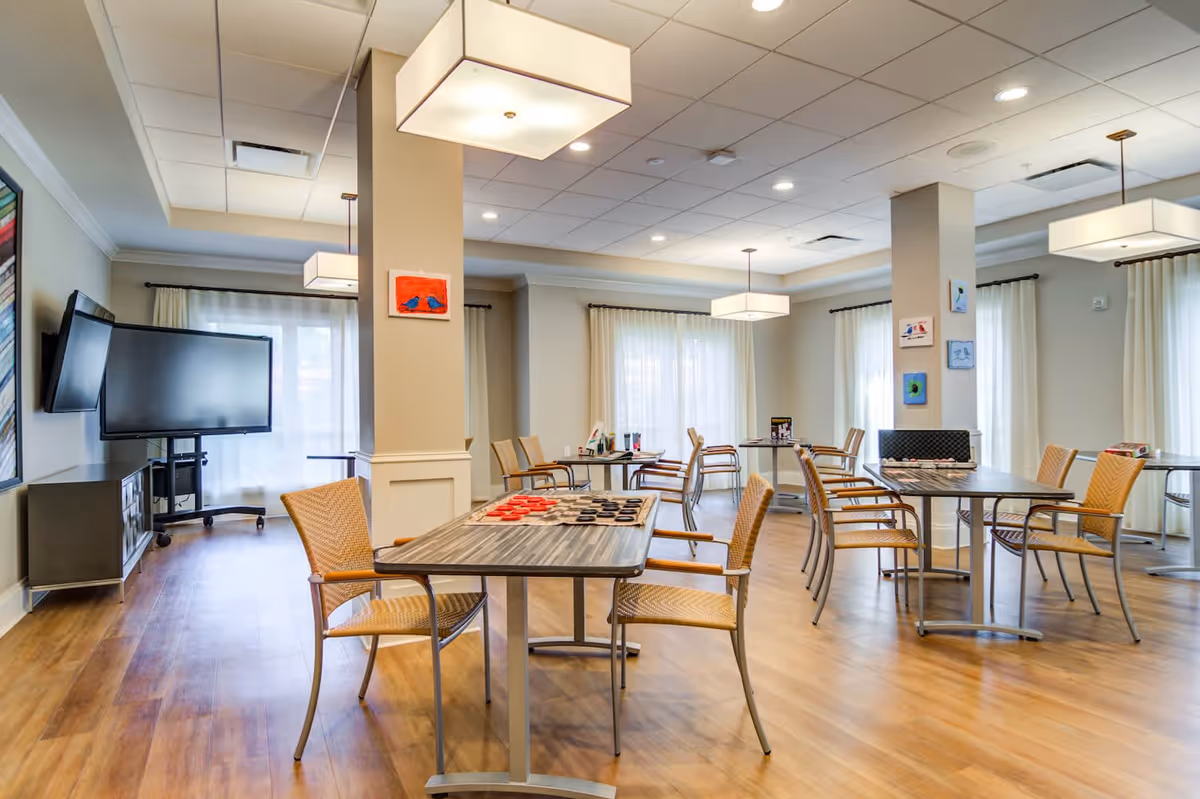 A bright and spacious common area with multiple tables and chairs arranged for group activities. The room features large windows with sheer white curtains, modern ceiling lights, and a flat-screen TV mounted on a stand. Some tables have board games set up, and the walls are decorated with small colorful paintings.