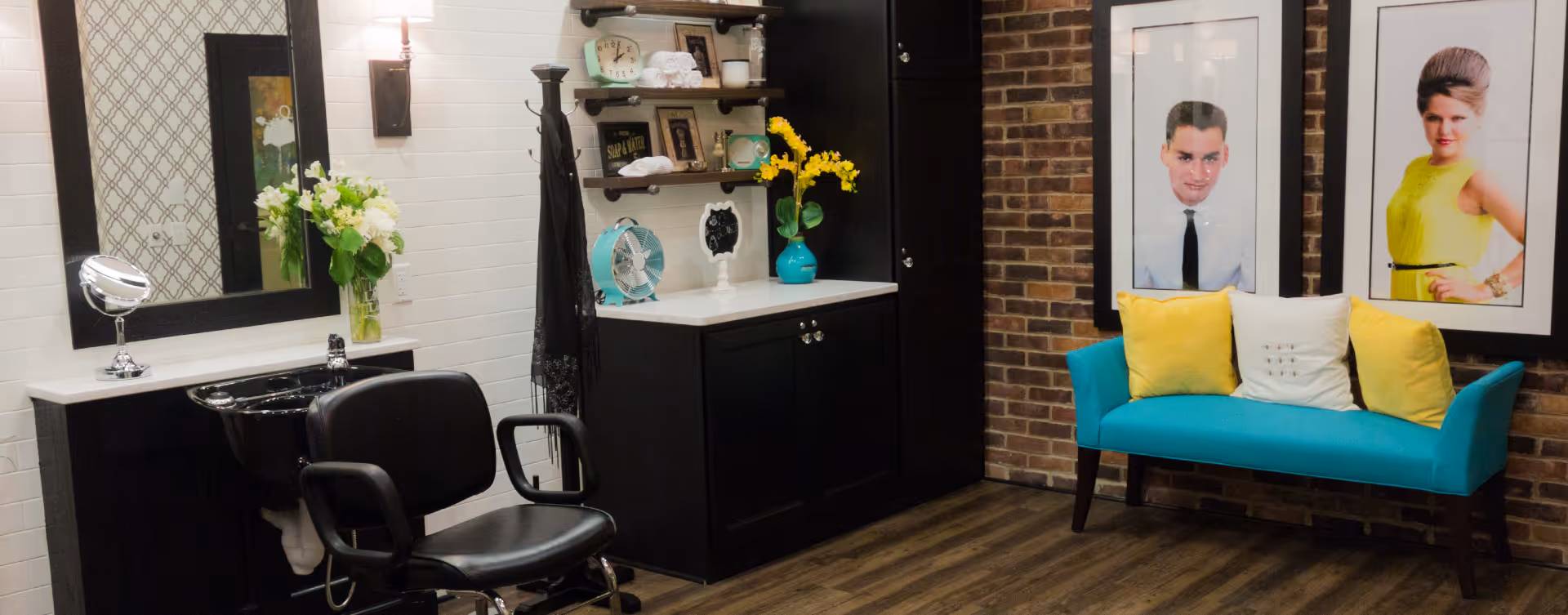 Interior of a stylish salon area with a black salon chair and sink on the left, a countertop with shelves holding decorative items and flowers, and a turquoise loveseat with yellow and white pillows against a brick wall with two framed portraits.