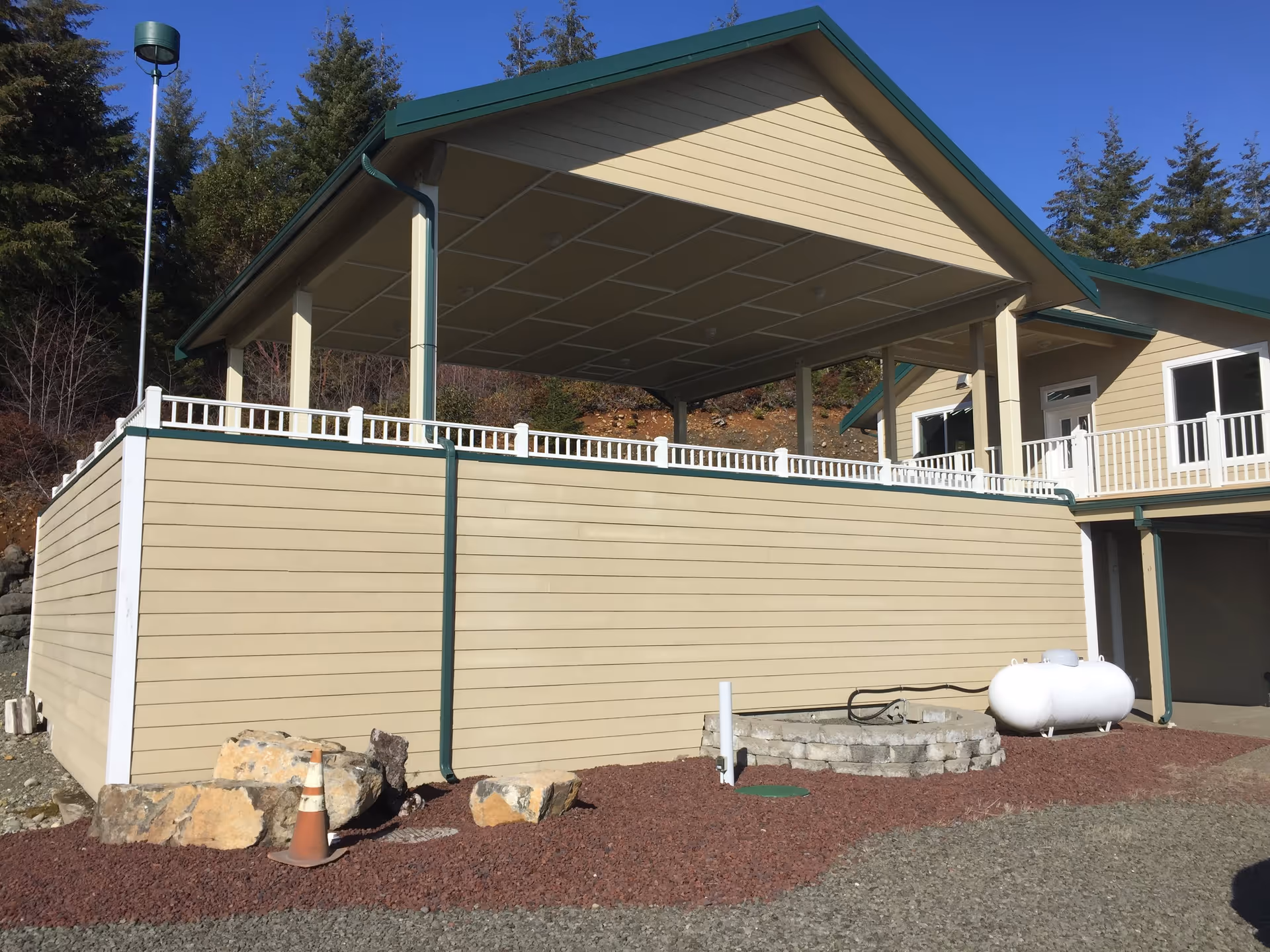 Exterior view of a beige building with green trim, featuring a covered patio area with white railing. The ground is covered with reddish gravel and large rocks, and there is a white propane tank and a small stone fire pit near the building. Trees and a clear blue sky are visible in the background.