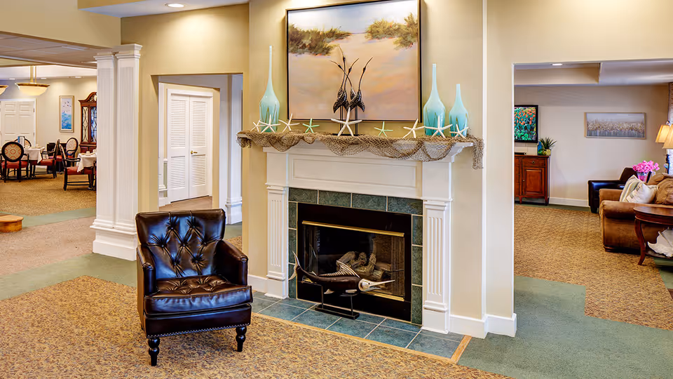 A cozy interior space featuring a white fireplace with green tile accents, decorated with starfish, fishing net, and two tall blue vases. Above the fireplace is a painting of a sandy beach scene with grasses. In front of the fireplace is a dark brown leather armchair. To the left, there is a dining area with tables and chairs, and to the right, a sitting area with brown sofas and a side table with a lamp and flowers.