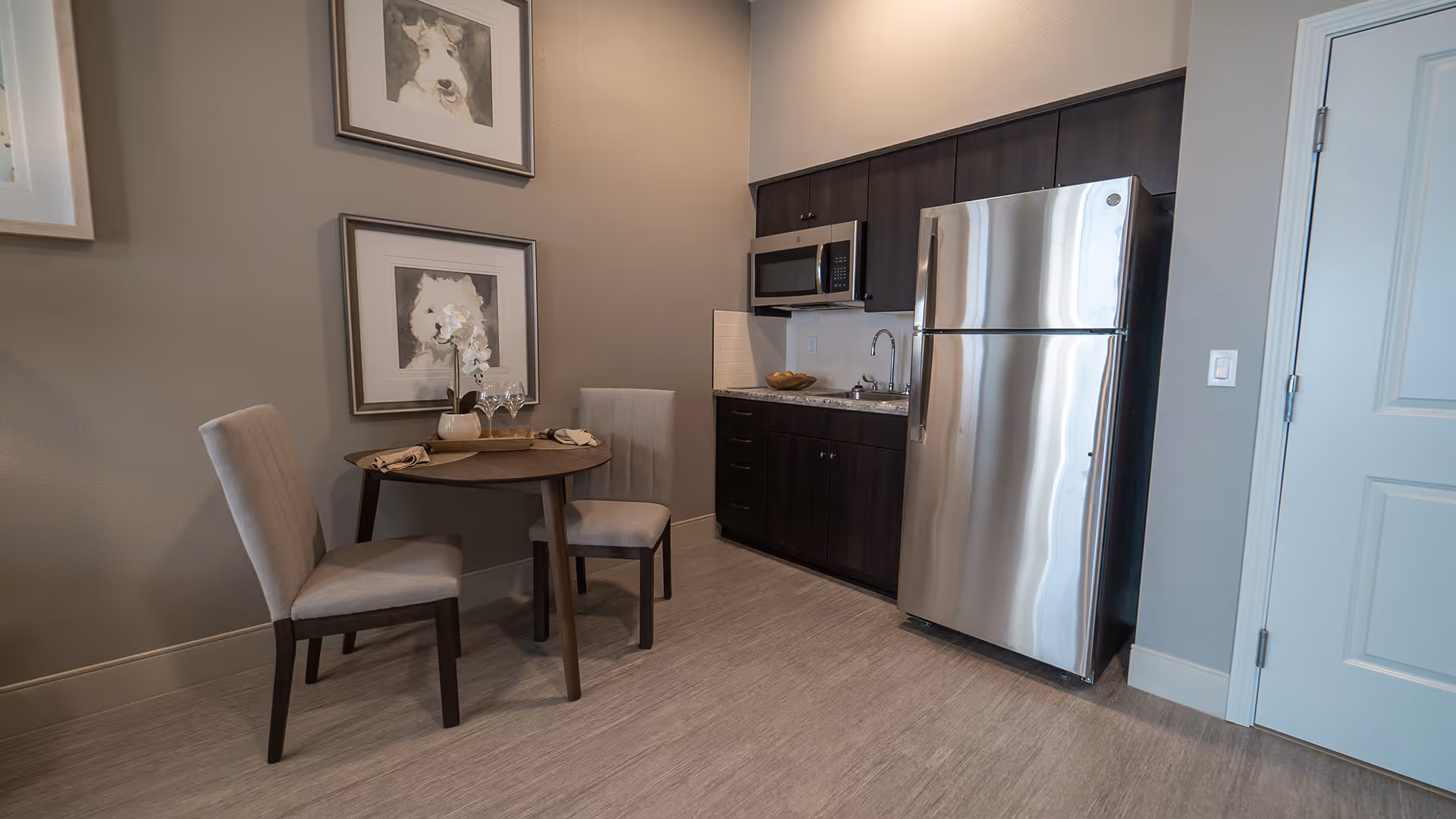 Small kitchenette with a stainless steel refrigerator, microwave and sink next to a round dining table with two chairs and framed wall art.