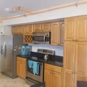 A kitchen with wooden cabinets, a stainless steel refrigerator, microwave, and stove. There is a black countertop with a kettle on the stove and a blue towel hanging from the oven handle. A wine rack is built into the cabinetry.