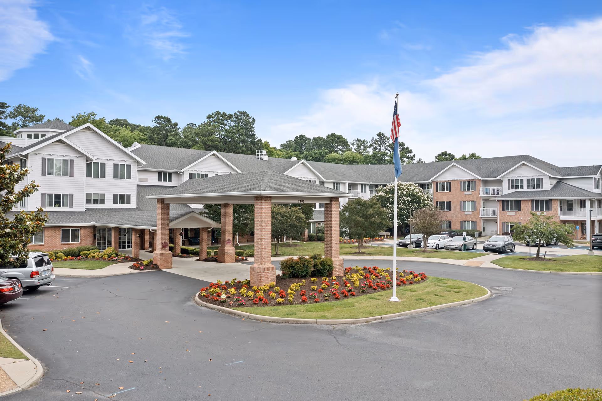 Exterior view of Colonial Harbor Gracious Retirement Living facility showing a large multi-story building with a covered entrance, landscaped flower beds, a flagpole with the American flag, parked cars, and trees in the background under a partly cloudy sky.