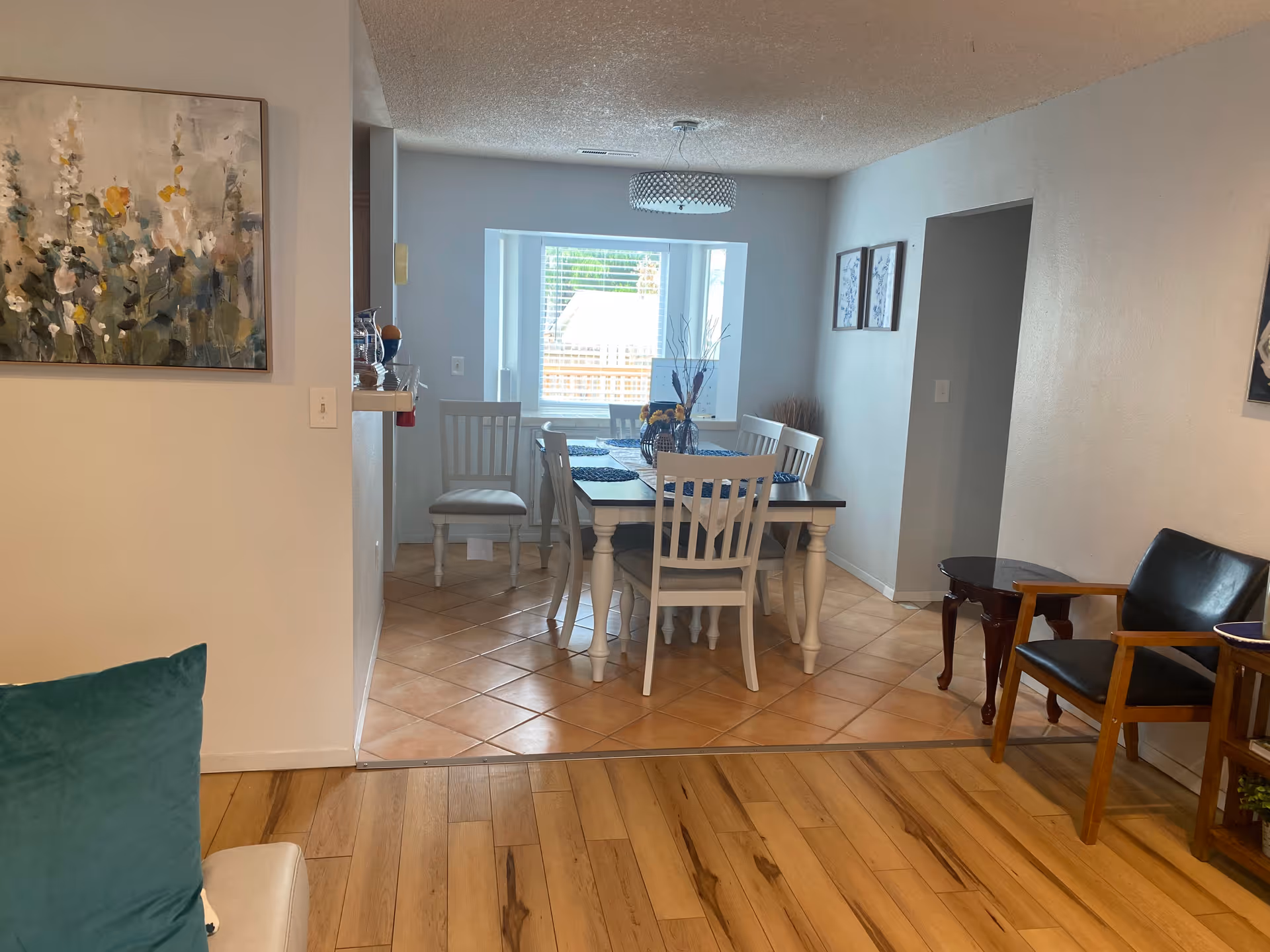 View of a dining area with a rectangular wooden table and six white chairs. The floor is tiled in the dining area and wooden in the foreground. There is a window with blinds at the far end, a modern ceiling light fixture above the table, and decorative artwork on the walls. To the right, there is a black leather chair and a small wooden side table.