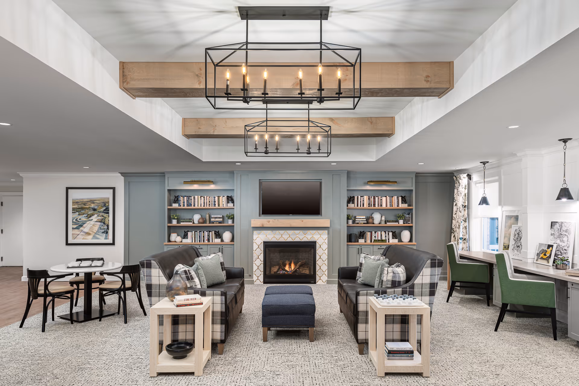 A cozy living room area with two plaid armchairs and a dark leather sofa arranged around a central ottoman. There is a fireplace with a TV mounted above it, flanked by built-in bookshelves filled with books and decorative items. The ceiling features wooden beams and two modern rectangular chandeliers. To the right, there is a desk area with green chairs and pendant lights, and to the left, a small round table with four chairs. The room has a neutral color palette with light walls and carpeted flooring.