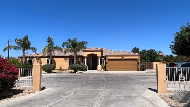 Single-story stucco building with an attached garage, gated concrete driveway, palm trees, and clear blue sky.