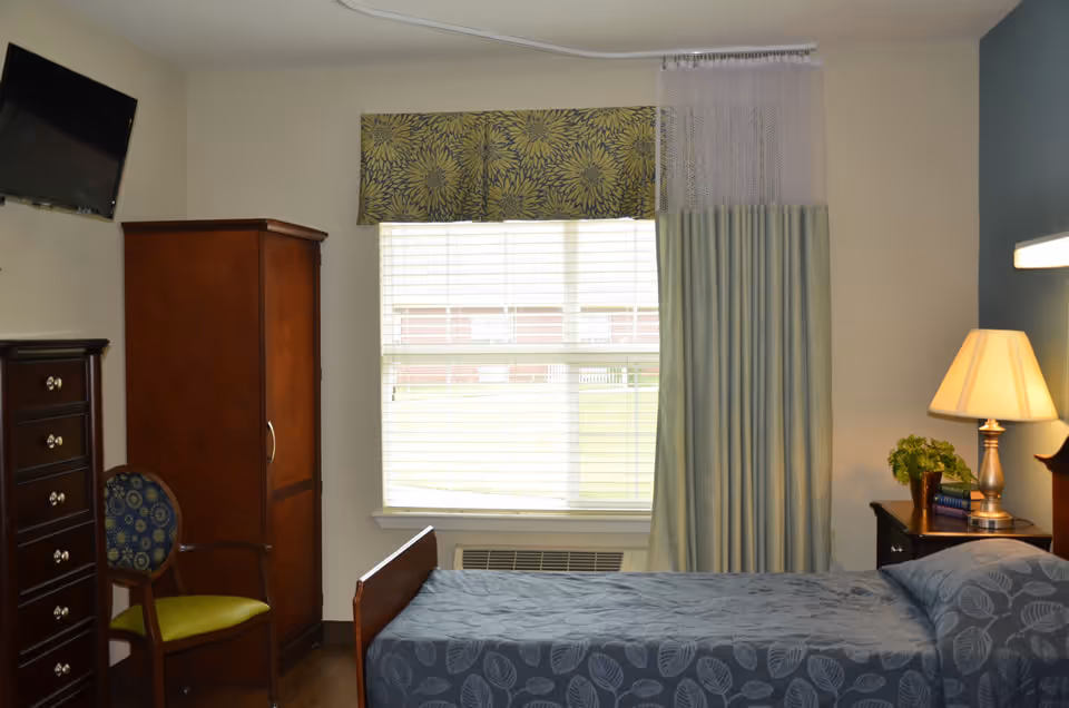 A senior care facility bedroom with a single bed covered in a blue leaf-patterned bedspread. Next to the bed is a nightstand with a lamp, a small plant, and some books. There is a large window with blinds and a floral valance, partially covered by a privacy curtain. A wooden wardrobe, a chest of drawers, and a chair with a green seat cushion and patterned backrest are also visible. A wall-mounted TV is positioned above the chest of drawers.