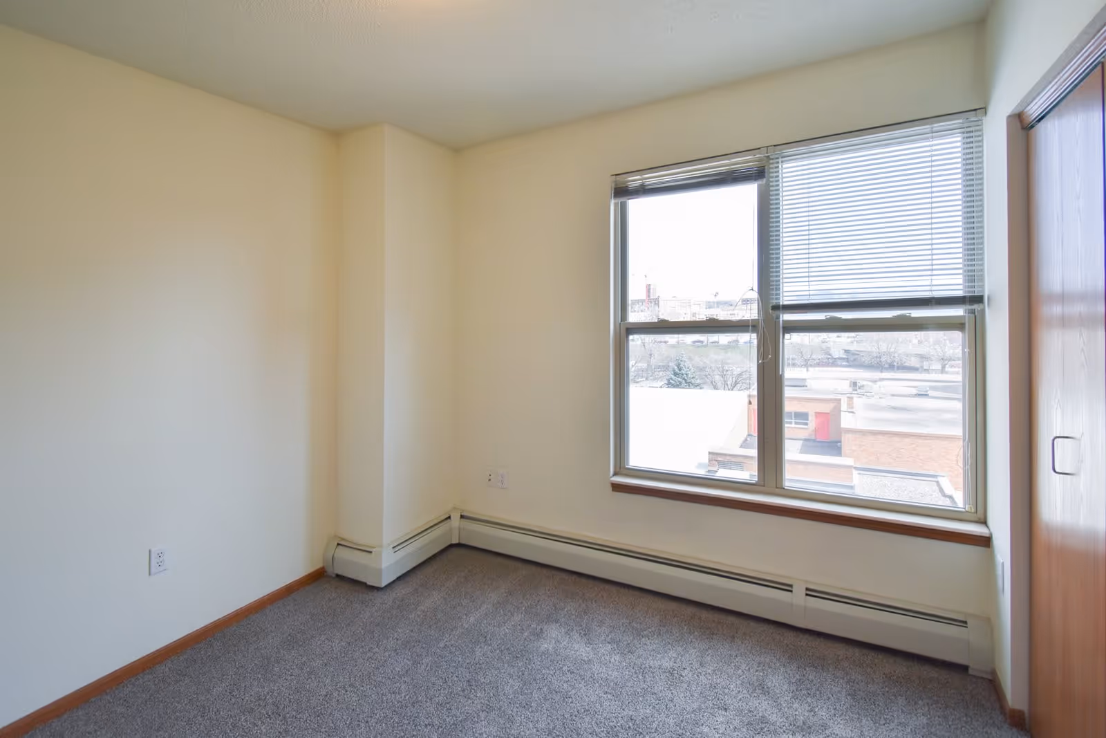 Empty small room with beige walls, gray carpet, a large window with blinds partially open showing an outside view, and a wooden closet door on the right side.