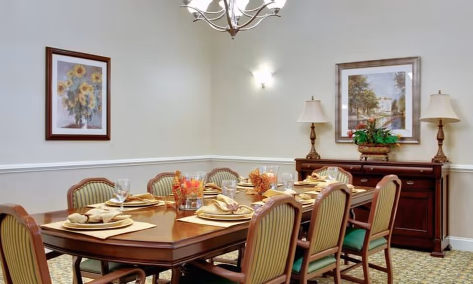 A formal dining room with a large wooden dining table set for eight people. The table has place settings with plates, napkins, and glasses. There are decorative centerpieces with flowers and candles. The room features two framed paintings on the walls, two table lamps on a wooden sideboard, and a chandelier hanging from the ceiling.