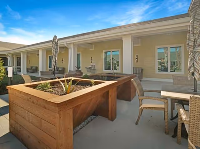 Outdoor patio courtyard with raised wooden planters, tables, chairs, umbrellas, and a covered walkway along the building.