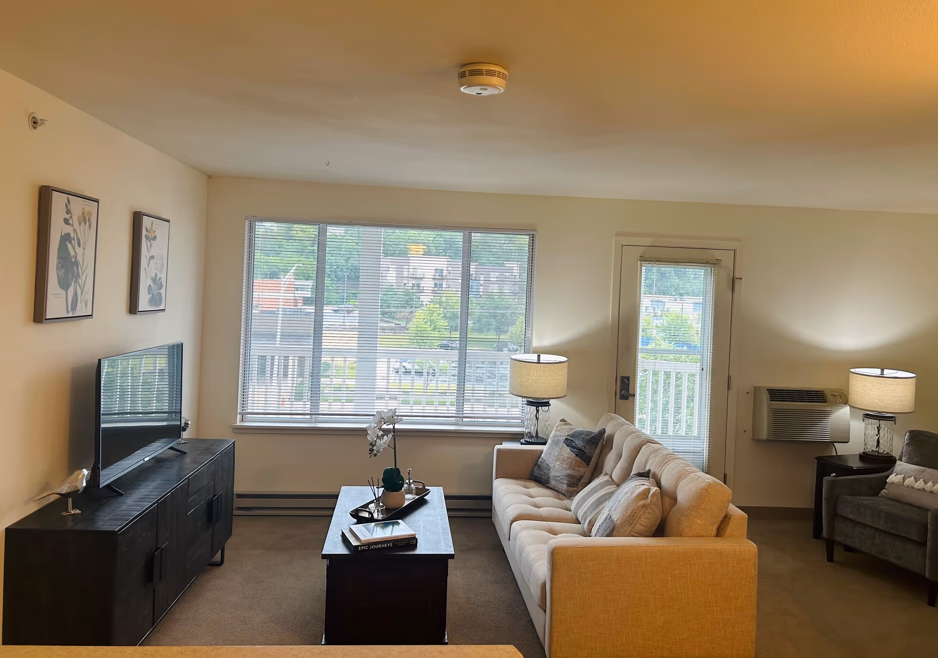 Furnished senior living room with a beige sofa, coffee table, TV on a console, armchair, lamps, and a large window and door to a balcony.