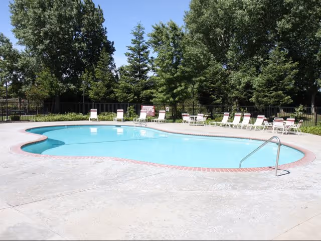 A kidney-shaped outdoor swimming pool with lounge chairs arranged along the deck and trees in the background.