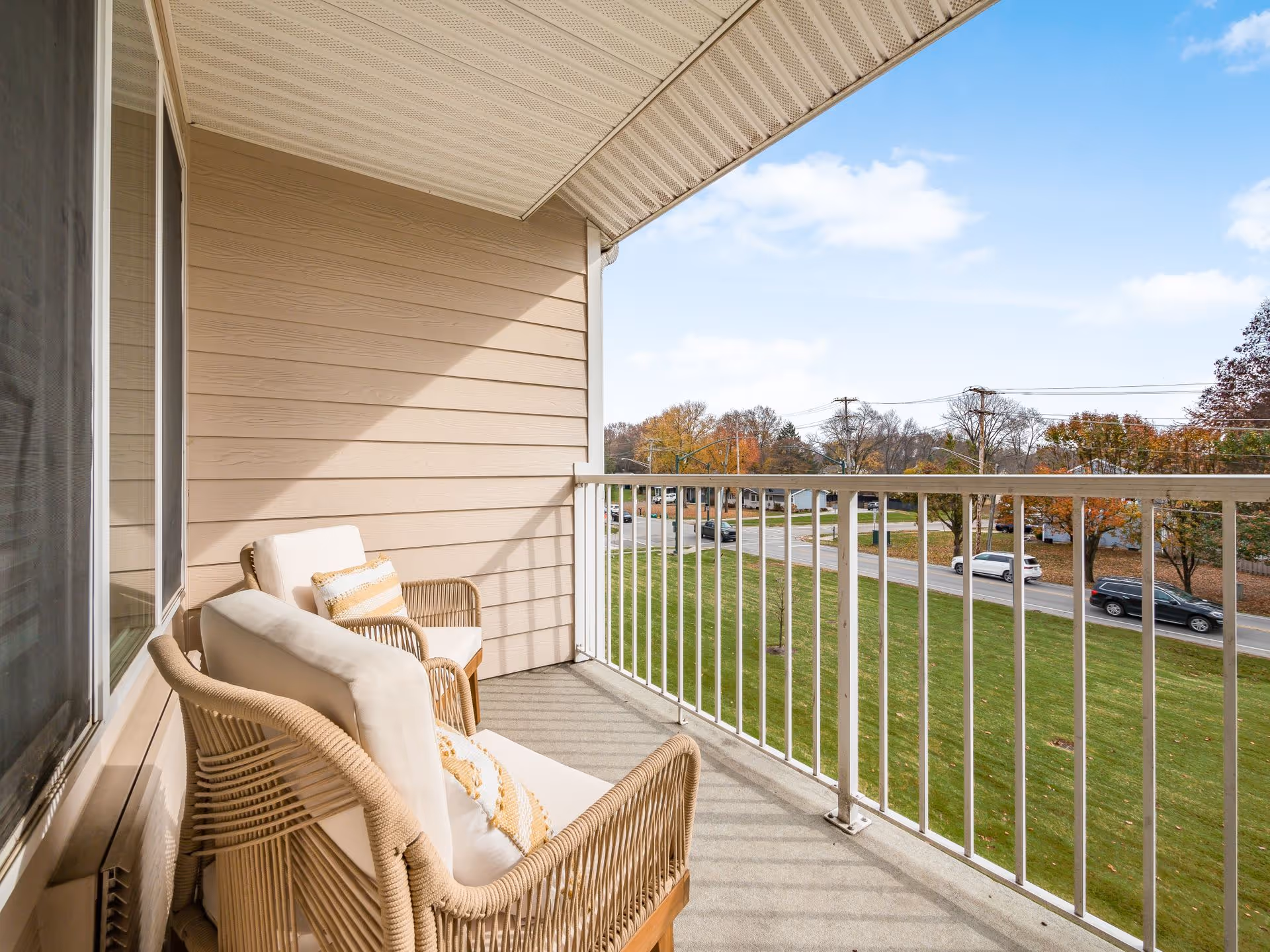 A balcony with two cushioned wicker chairs and a small pillow on each chair. The balcony has a white railing and overlooks a grassy area with a road and several cars. Trees with autumn foliage are visible in the background under a partly cloudy sky.