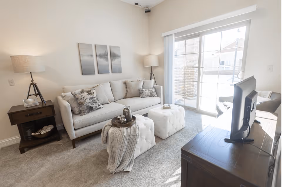 A bright living room with a light gray sofa adorned with patterned cushions, two white tufted ottomans, a wooden side table with a lamp, and a wooden TV stand with a flat-screen TV. The room has a large sliding glass door leading to an outdoor patio, allowing natural light to fill the space. Three framed landscape pictures hang on the wall above the sofa.