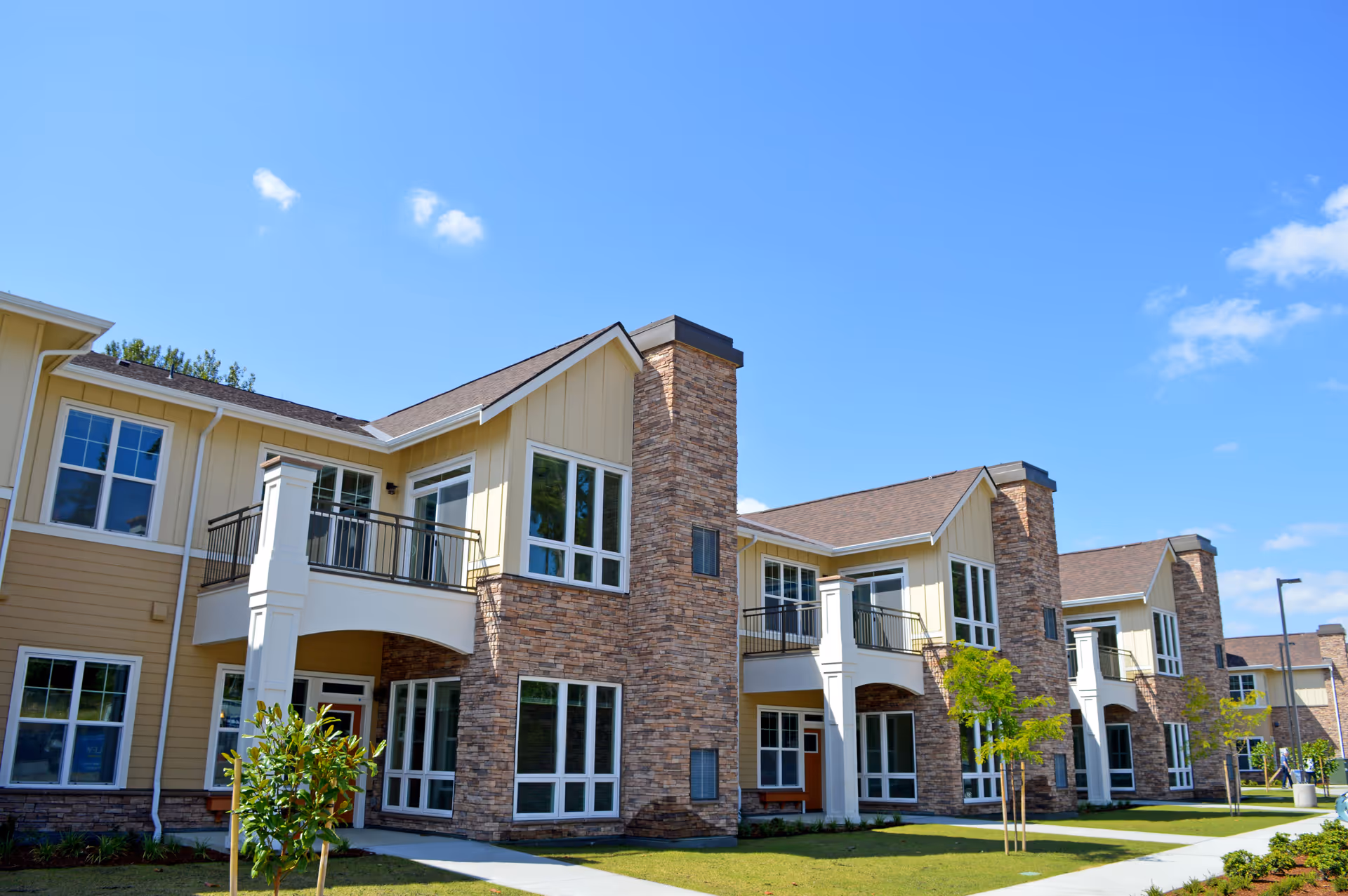 Exterior view of a modern senior living facility building with beige siding and stone accents, featuring multiple windows, balconies, and a well-maintained lawn under a clear blue sky.