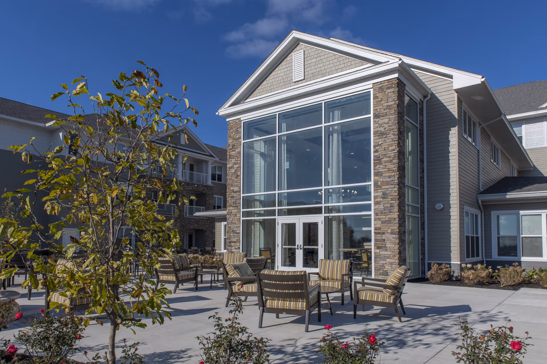 Outdoor patio area of a senior living facility with several striped cushioned chairs and small tables arranged on a concrete surface. The building features large glass windows with stone and siding exterior under a clear blue sky. There are some small trees and flowering plants around the patio.