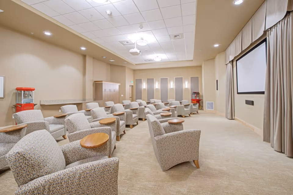 Small screening room with rows of upholstered armchairs each with wooden side tables facing a wall-mounted projection screen.