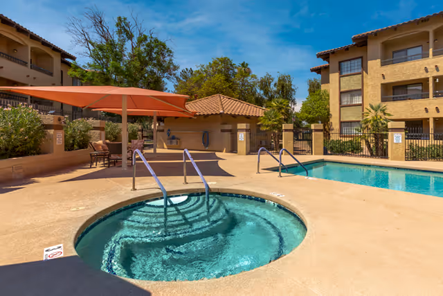 Outdoor pool area at Brookdale Springs Mesa featuring a round hot tub in the foreground with metal handrails, a rectangular swimming pool in the background, shaded seating area with an orange canopy, and multi-story residential buildings surrounded by trees under a clear blue sky.