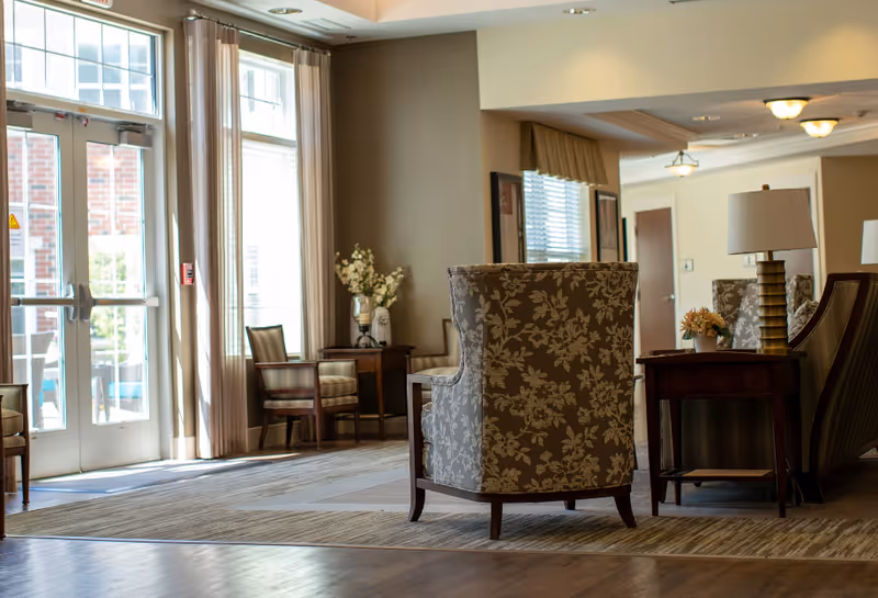 A cozy seating area in a senior living facility with floral patterned armchairs, wooden side tables with lamps and flower arrangements, large windows with sheer curtains allowing natural light, and a glass door leading outside.