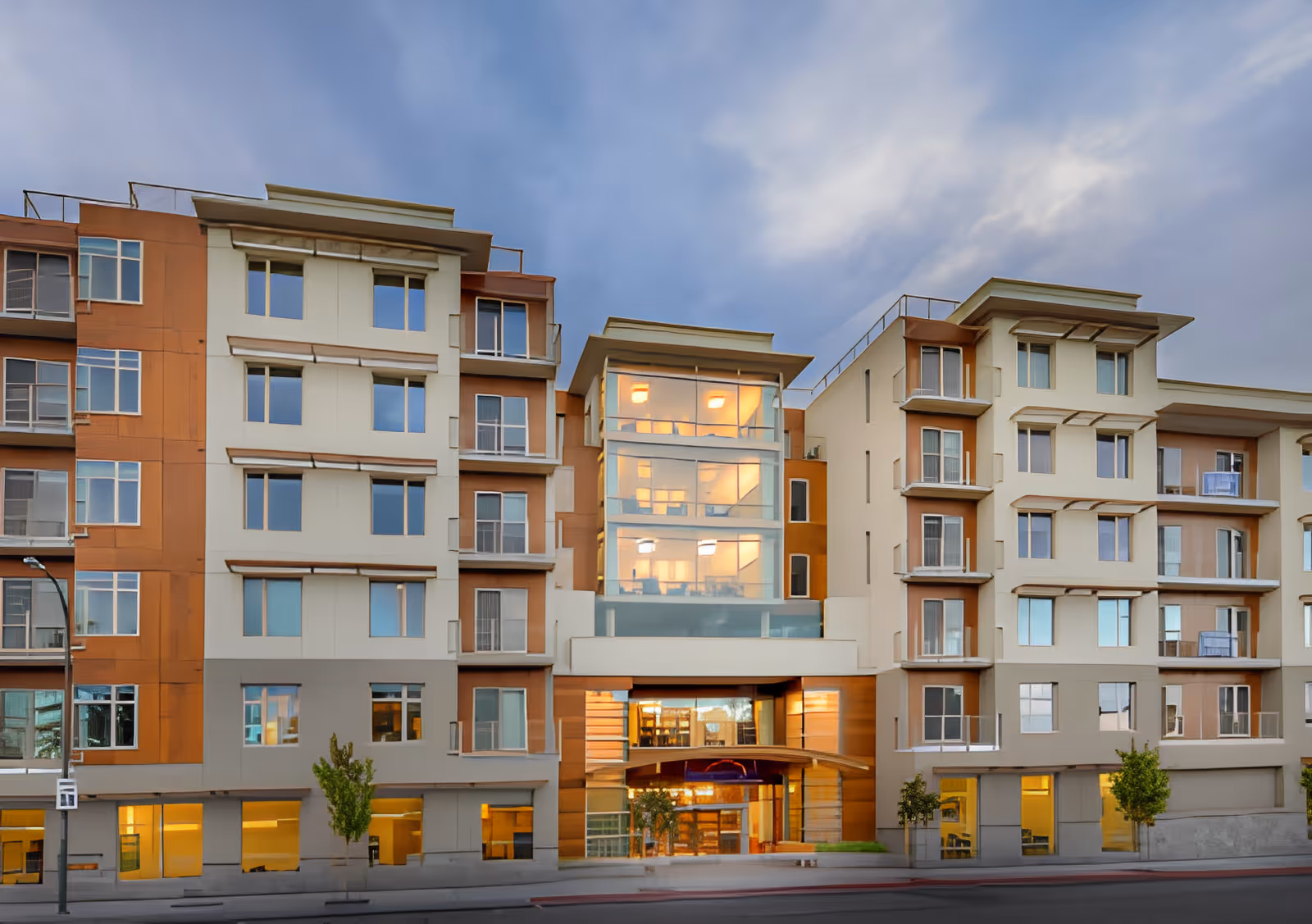 Modern multi-story senior living building front with a central glass atrium and balconies at dusk.