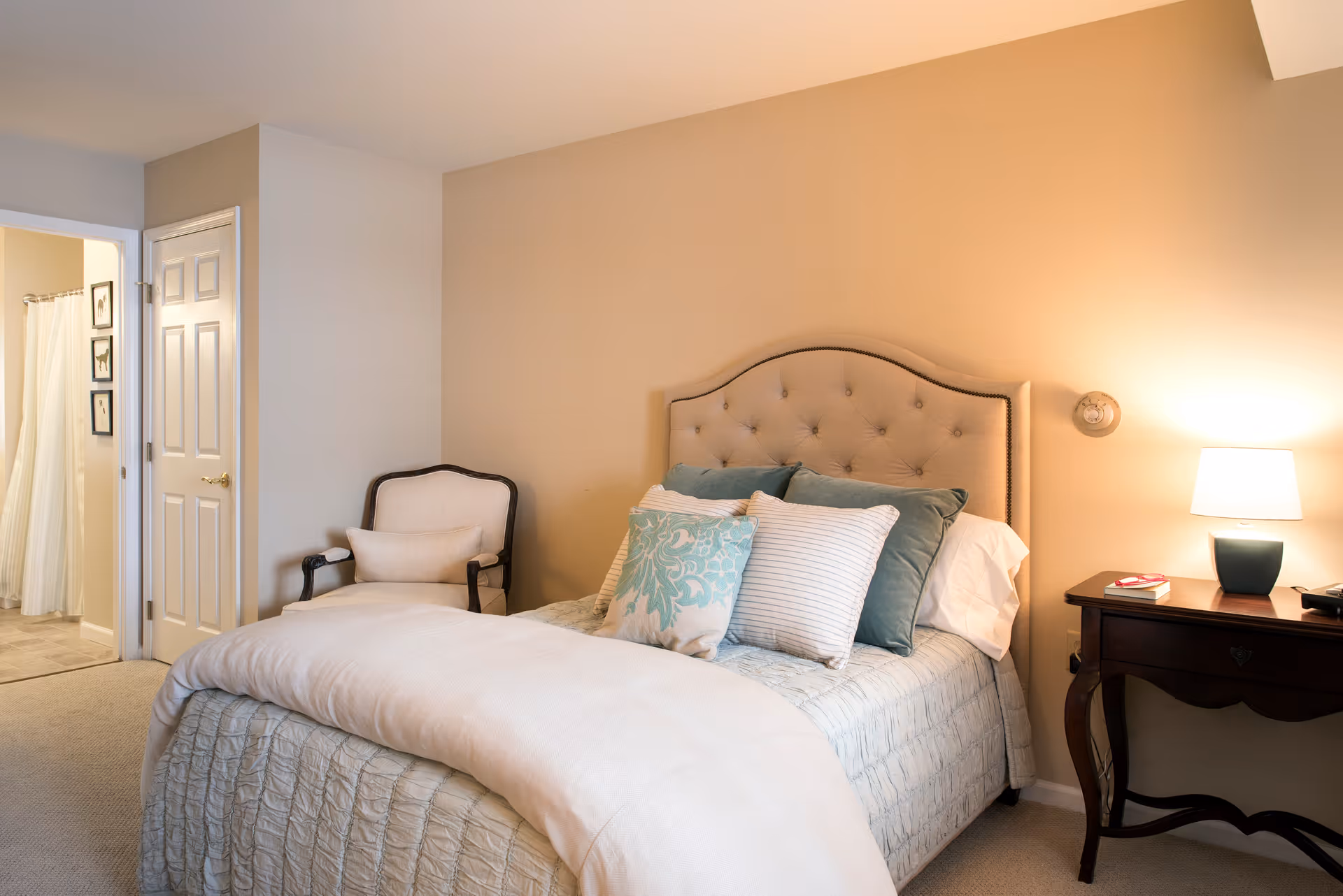 A cozy bedroom with a beige tufted headboard bed adorned with multiple pillows in shades of blue and white. To the right of the bed is a wooden nightstand with a lit lamp and a book. To the left, there is an upholstered armchair with a cushion. In the background, an open door reveals a bathroom with a shower curtain and framed pictures on the wall.