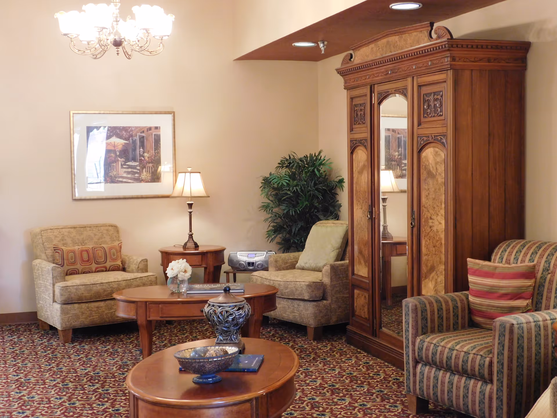 A cozy living room area with patterned carpet, a chandelier, two upholstered armchairs with cushions, a wooden coffee table with decorative items, a wooden armoire with mirrored doors, a side table with a lamp, a potted plant, and a framed picture on the wall.