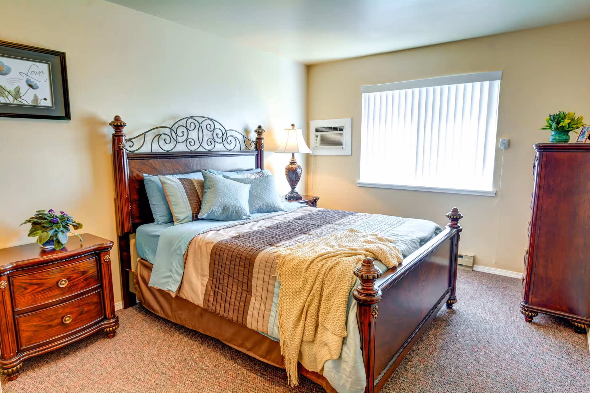 A well-lit bedroom featuring a wooden bed with an ornate headboard, neatly made with blue and brown bedding and several pillows. There is a wooden nightstand with a lamp and a small plant on the left side of the bed, a window with vertical blinds on the right wall, and a wooden dresser with a plant and framed photo on top. The room has beige walls and carpeted flooring.