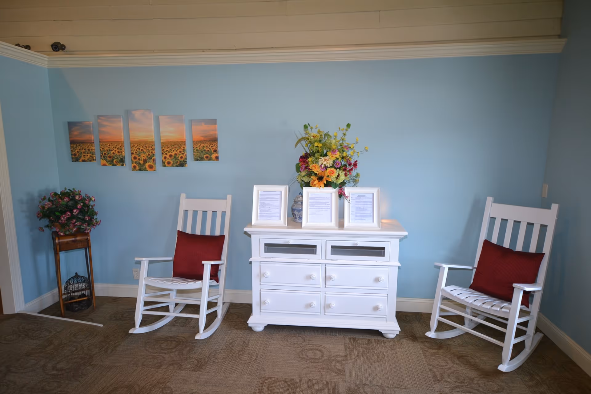 A cozy sitting area with two white wooden rocking chairs each with a red cushion, a white cabinet with drawers in the center, and a vase of colorful flowers on top. On the light blue wall behind, there is a multi-panel artwork depicting a sunflower field at sunset. A small wooden stand with a potted plant is placed to the left of the left rocking chair.